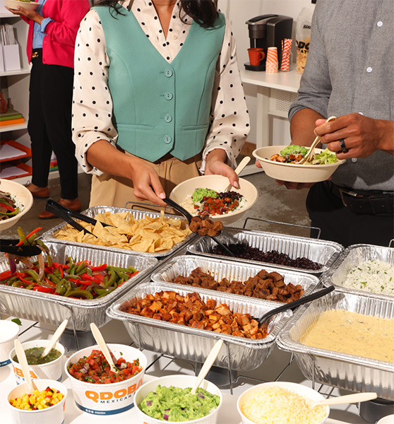 People serving themselves from a QDOBA catering hot bar featuring a variety of proteins, toppings, and sides, suitable for business occasions like office lunches, conferences, and company retreats.