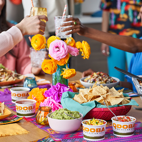 People toasting with drinks around a colorful table set with QDOBA catering items, including chips, guacamole, salsa, and vibrant floral decorations.