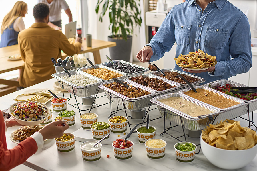 Employees serving themselves from a QDOBA customizable hot bar featuring trays of various proteins, toppings, and sides, with an office setting and people working in the background.