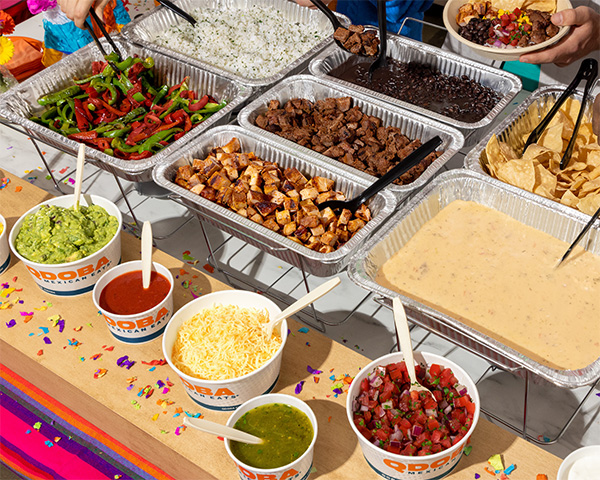 QDOBA Catering custom Hot Bar setup with trays of grilled vegetables, cilantro lime rice, black beans, steak, chicken, queso, chips, guacamole, salsa, shredded cheese, and other toppings, decorated with colorful confetti.