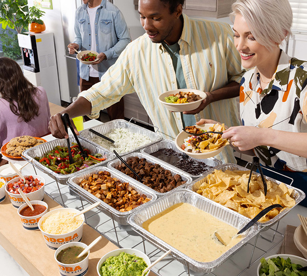 QDOBA Catering Hot Bar setup in an office environment with trays of rice, beans, meat, and various toppings, as well as people serving themselves and enjoying the meal in the background.