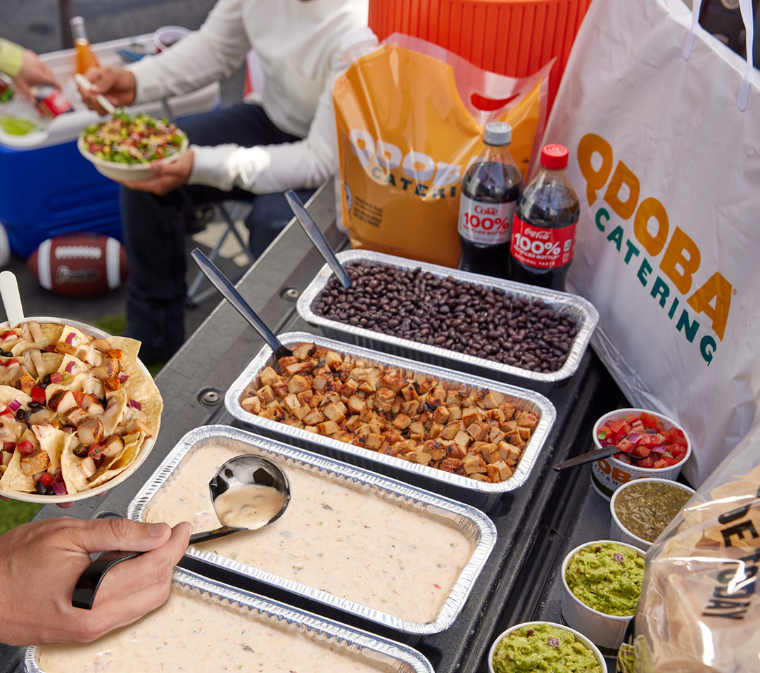 A QDOBA Catering Customizable Hot Bar at a football game tailgate party. Pans of beans, Adobo-seasoned chicken, Three Cheese Queso, guacamole, and a variety of salsas and toppings are displayed on the open back of a pickup truck. Behind the food, there is a large QDOBA Catering bag. In the bottom left corner, a hand is scooping Three-Cheese queso onto a bowl of nachos. In the top left corner, someone is sitting next to an open cooler of soda bottles, eating a burrito bowl, with an American Football by their feet. Another person's arm reaches in to grab a bottle from the cooler.