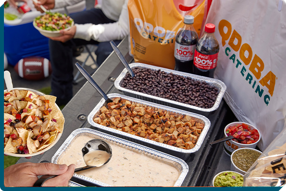 QDOBA's game day catering display with a tailgate pack featuring tortilla chips, savory queso dip, fresh toppings, and a nacho bar setup, ready to fuel fans at a football party.