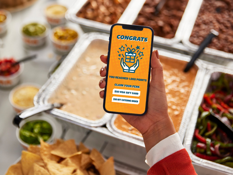 Hand holding a smartphone showing QDOBA rewards screen with a catering setup in the background, displaying options to claim a $50 Visa gift card or $50 off a catering order.