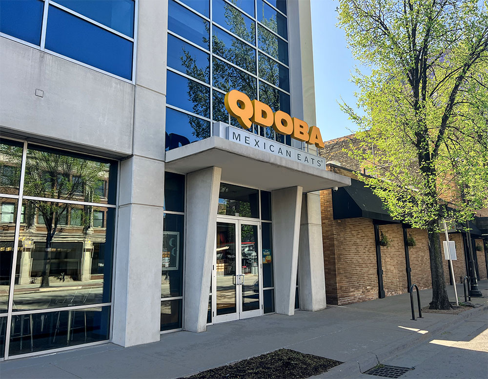 Exterior photography of the QDOBA Mexican Eats restaurant located on Water Street in downtown Milwaukee, near the Fiserv Forum and at the edge of the RNC security zone. The weather is sunny and the sidewalk in front of the restaurant is shaded by a tree.