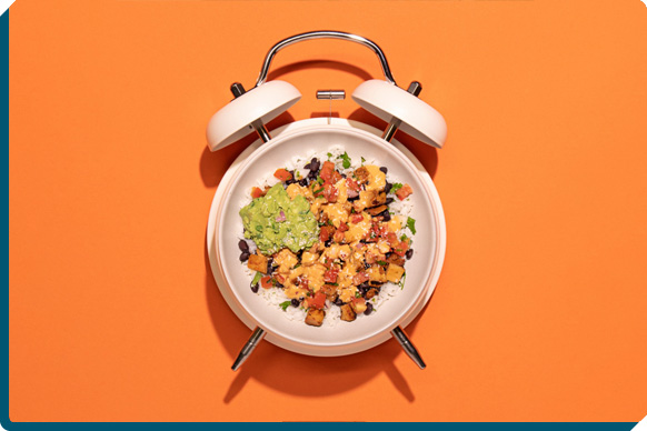 QDOBA game day burrito bowl served on a white plate with alarm clock hands, symbolizing quick and easy party planning for game day festivities, featuring seasoned adobo chicken, savory black beans, spicy Queso Diablo, freshly made guacamole, and pico de gallo.