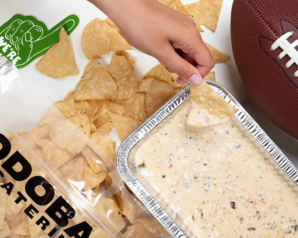 Image: A hand dipping a tortilla chip into a pan of QDOBA Mexican Eats' signature Three-Cheese Queso. Freshly-made tortilla chips scattered on the table. An American football partially visible in the top right corner. To the left, a green foam finger that says 'We're #1.' Perfect appetizer for QDOBA Catering and game day watch party.