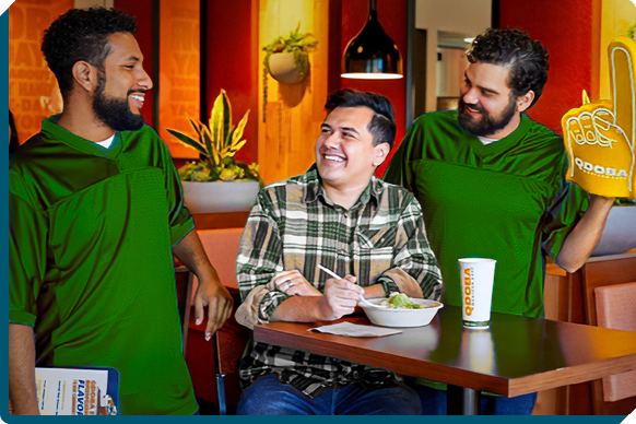 Enthusiastic friends in green and gold gear enjoying a laugh over a QDOBA Mexican Eats meal at a football watch party, representing a mix of game day excitement and casual dining with company.