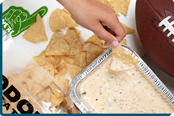 A hand dipping a golden tortilla chip into QDOBA's rich and creamy three-cheese queso next to a vibrant football party setup, showcasing the ease of game day catering with freshly prepared snacks.