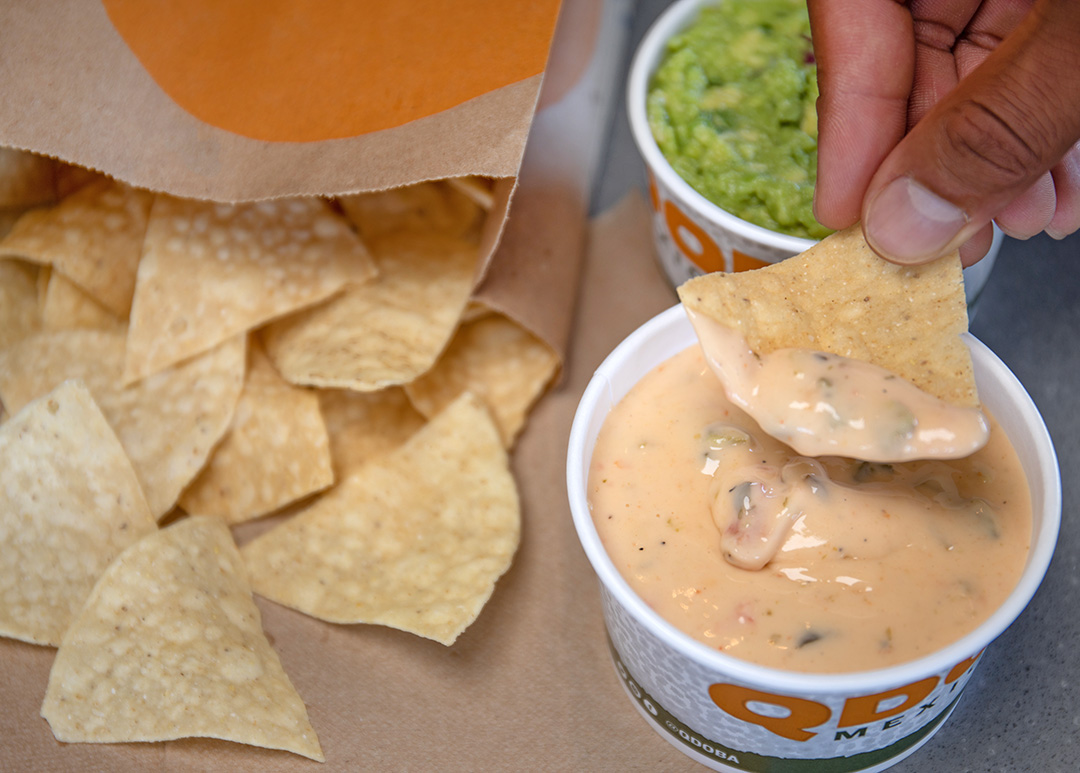 Image: Indulge in the spirit of game day with QDOBA Chips & Queso! In the top right corner, fingers holding a tortilla chip dip into a cup of QDOBA's signature Three-Cheese Queso. Behind the cup of queso is a cup of fresh hand-smashed guacamole. On the left half, tortilla chips spill out from the top of a QDOBA takeout paper bag. Enjoy this flavor-packed treat during the Green & Gold Fridays promotion and celebrate your love for the Green Bay Packers in style! 