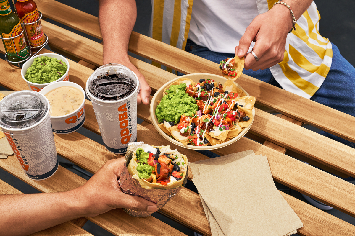 Outdoor table with QDOBA Mexican Eats food, including a burrito, nachos topped with fresh ingredients, guacamole, queso dip, and beverages, perfect for a quick bite during the RNC in Milwaukee.