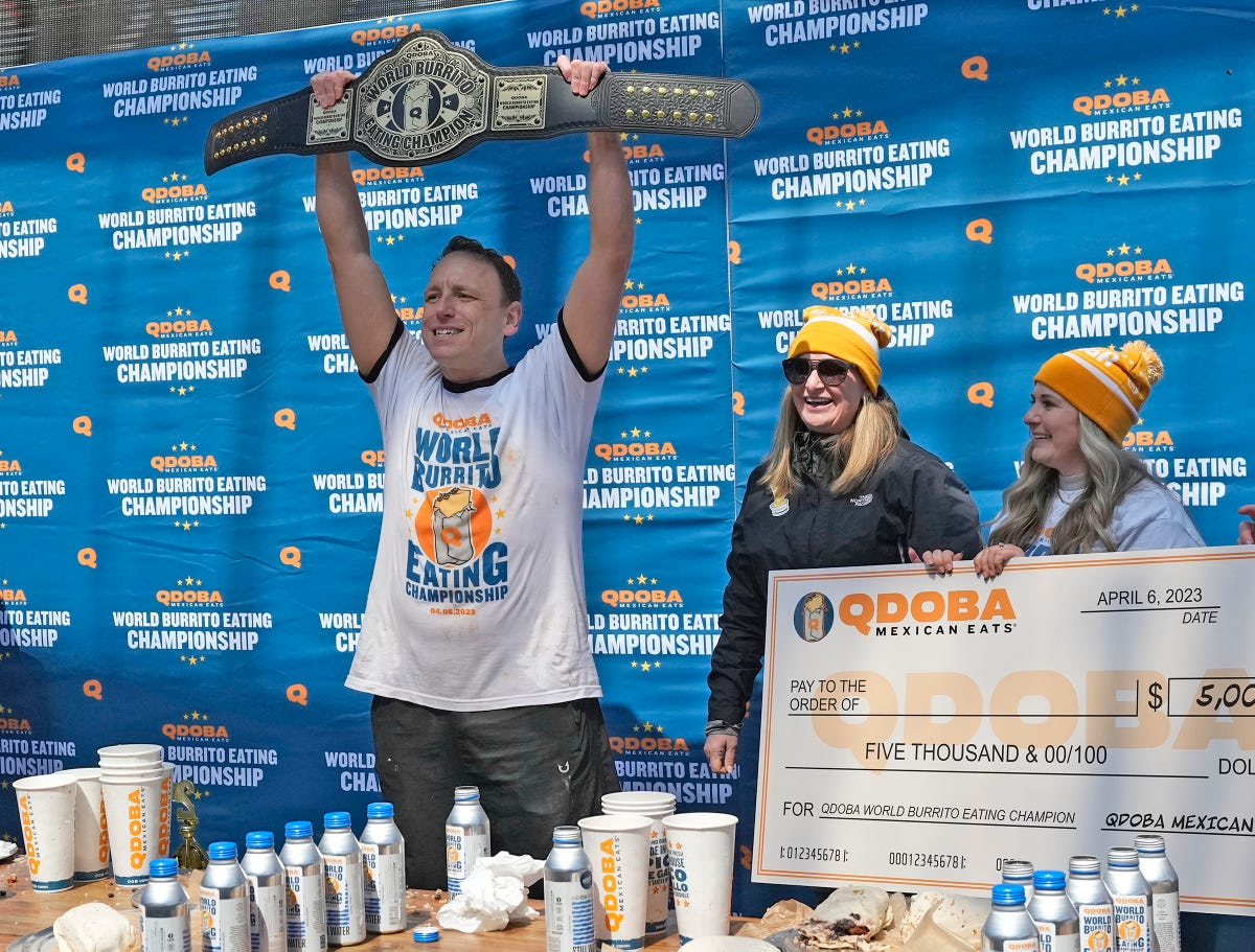 Winner Joey Chestnut holding a championship belt and a $5,000 check at the QDOBA World Burrito Eating Championship, with two women in QDOBA gear congratulating him, set against an event-branded backdrop in Milwaukee’s Deer District in 2023.