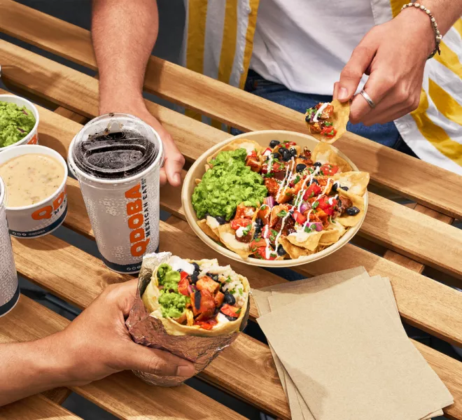 Outdoor table with QDOBA Mexican Eats food, including a burrito, nachos topped with fresh ingredients, guacamole, queso dip, and beverages, perfect for a quick bite during the RNC in Milwaukee.