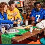 Excited friends sharing easy football party food from QDOBA Mexican Eats, with colorful football jerseys and a festive atmosphere, embodying fun football party ideas and snacks.