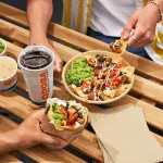 Outdoor table with QDOBA Mexican Eats food, including a burrito, nachos topped with fresh ingredients, guacamole, queso dip, and beverages, perfect for a quick bite during the RNC in Milwaukee.